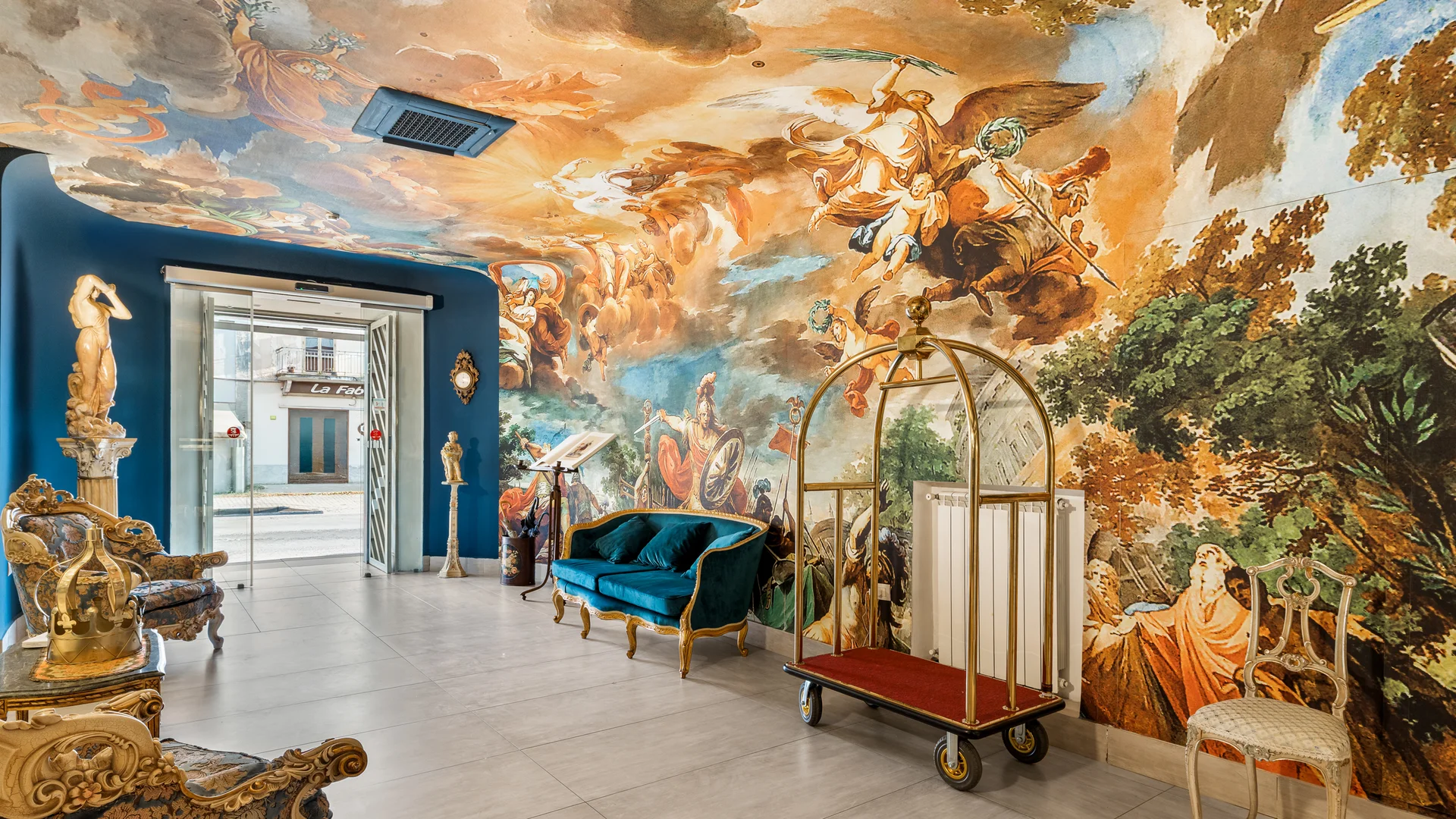 Hotel vicino alla Reggia di Caserta · Regina 4★ Interno di hotel con pareti affrescate, divano blu e carrello portabagagli dorato