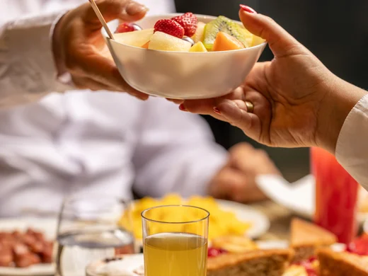 Hotel vicino alla Reggia di Caserta · Regina 4★ Due persone che si passano una ciotola di frutta fresca a una tavola con pane e succo