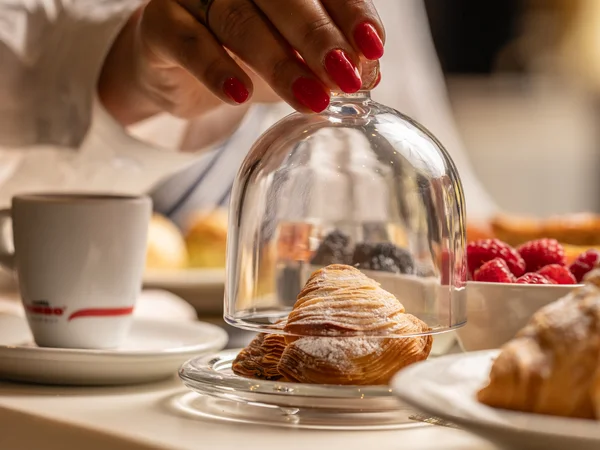 Hotel vicino alla Reggia di Caserta · Regina 4★ Mano con unghie rosse che solleva una campana di vetro su un croissant