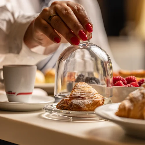 Hotel vicino alla Reggia di Caserta · Regina 4★ Mano con unghie rosse che solleva una campana di vetro su un croissant